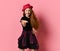 Teen female in black dress, checkered shirt, red hat. Smiling, reaching for you by her hand, posing on pink background. Close up