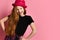 Teen female in black dress, checkered shirt on waist, red hat. She is laughing, hands on hips, posing on pink background. Close up