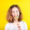 Teen girl angry bites a multi-colored round lollipop on yellow background