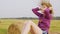 Teen girl puts straw wreath oh her head playing sitting on hay stack, side view.