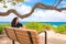 Teen girl sitting on bench under trees by ocean