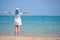 Teen girl in white dress and hat standing barefooted with wide spread arms on beach enjoying tropic vacations looking at