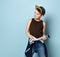 Teenage boy in cap, brown undershirt and jeans. He is tying hoodie around his waist and smiling. Posing on blue studio background