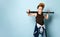 Teenage boy in cap, undershirt, jeans and hoodie tied around his waist. Holding black baseball bat, posing on blue background