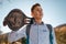 A teenage boy holds a skateboard in his hands and looks away. Mountains and sky in the background. Bottom view