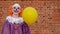 A teenage boy in a scary clown costume with teeth smiles funny against a red brick wall. Yellow balloon. Street makeup on the face