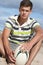 Teenage Boy Sitting On Beach Holding Rugby Ball