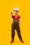 Teenage boy in work jumpsuit and protective helmet holds drill. Child is like worker builder on yellow background