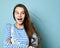 Teenage female in jeans overall, striped sweatshirt. She smiling, showing thumbs up, winking, posing on blue background. Close up