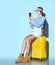 Teenage female in overall, sweatshirt, boots, sun visor cap. She using smartphone, sitting on yellow suitcase, blue background.