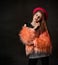 Teenage girl with african pigtails, in red hat, orange faux fur coat. Propping her chin by forefinger, posing on dark background