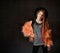 Teenage girl with african pigtails, in red hat, orange faux fur coat. Propping her chin by forefinger, posing on dark background