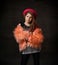 Teenage girl with african pigtails, in red hat, orange faux fur coat. Propping her chin by forefinger, posing on dark background