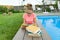 Teenage girl in glasses reads a book, background swimming pool, lawn near the house. School, education, knowledge, adolescents.