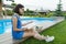 Teenage girl in glasses reads a book, background swimming pool, lawn near the house. School, education, knowledge, adolescents.