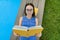 Teenage girl in glasses reads a book, background swimming pool, lawn near the house. School, education, knowledge, adolescents.