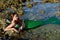 A teenage girl in a green mermaid costume lies in the water among the stones.