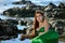 The teenage girl in the green shiny mermaid costume lies at sunset in the water among the stones on the coast and looks to the sid