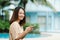 A teenage girl with long hair Asian girl sits by the pool in a hotel with her cell phone and happily turning to the camera on a