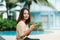 A teenage girl with long hair Asian girl sits by the pool in a hotel with her cell phone and happily turning to the camera on a