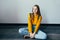 Teenage girl looking at camera and smiles sitting on wooden floor. Beautiful student girl in yellow sweater and blue jeans hold bo