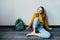 Teenage girl looking on cellphone and smiles sitting on wooden floor. Beautiful girl in yellow sweater and blue jeans with backpac