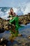 Teenage girl in a mermaid costume is sitting with her eyes closed on a rock on the beach against the backdrop of raging waves.