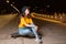 A teenage girl sits on a skateboard, posing on a night street. Outdoor. Side view. The concept of youth subculture