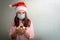 A teenage girl stands at home wearing a Santa hat and a medical mask, holding golden Christmas tree toys in her palms