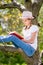 Teenage girl in white T-shirt and pink hat is sitting on a tree in the forest and reading great book. Beautiful idea and fun