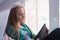 Teenage sits at the window and reading book in tablet