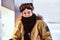 A teenage snowboarder dressed in snowsuit and protective goggles sitting on railing near the snowy beach