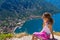 A teenage tourist girl looking down at the Boka Bay of Kotor from a high cliff sitting on a rock