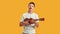 Teenaged disabled boy with Down syndrome having fun while playing ukulele, standing isolated over yellow background