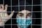 A teenager of Arabic appearance in a black hood and an individual mask stands behind bars, his hand on the bars.