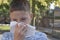 Teenager boy blows his nose in a paper handkerchief on a background of vegetation in a park. Sneezes