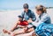 Teenager Boy passing drinking water bottle to Smiling Father in sunglasses with smartphone sitting with backpacks on the sandy