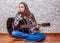 Teenager brunette girl with long hair sitting on the floor and playing an black acoustic guitar on gray wall background