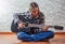 Teenager brunette girl with long hair sitting on the floor and playing an black acoustic guitar on gray wall background