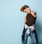 Teenager in cap, brown undershirt, jeans and hoodie tied around his waist. Smiling, showing thumb up, posing on blue background