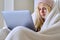 Teenager frozen in winter cold season, warming up with blanket hat, looking at laptop