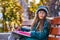 A teenager girl in glasses, a coat and a knitted hat sits on a bench in the park and draws a drawing on paper.