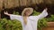 Teenager girl in hat and white dress standing with raised hands on summer plants background. Happy girl enjoying sunny