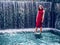 Teenager girl in high fashion red dress posing in water, water fall behind her back. Prom photoshoot in beautiful location Young