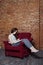 A teenager girl writes in a diary notebook sitting in an old chair on a gloomy abandoned station with a brick wall