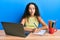 Teenager hispanic girl sitting on the table studying for school making fish face with mouth and squinting eyes, crazy and comical