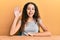 Teenager hispanic girl wearing casual clothes sitting on the table waiving saying hello happy and smiling, friendly welcome