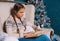 Teenager in a knitted sweater sits in an armchair by the Christmas tree and reads a book