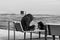 A teenager in a large hood sits on a bench by the sea and reads a book.