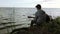 A teenager plays a guitar while sitting on the shore of a lake.